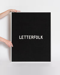 2 hands holding up the Letterfolk Black Wordsmith Letter Board against a grey backgeound. 