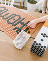 An image of a person using the Letterfolk Black tiles to design their Letterfolk Clay Tile Mat on a wooden table. 